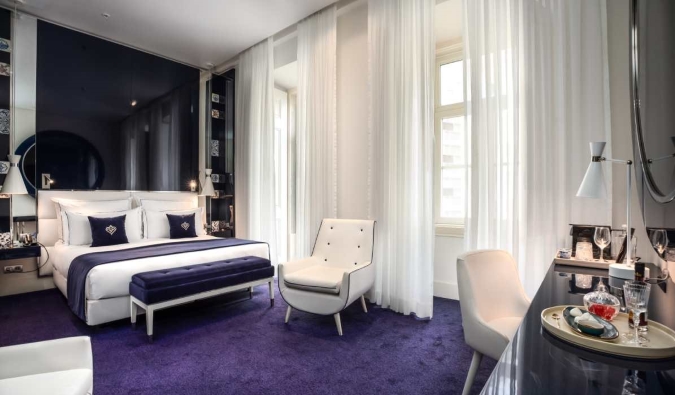 Një suitë luksoze me tapete blu të thellë, një tryezë elegante, karrige e bardhë, shtrat i gjerë dhe dritare franceze me perde të holla në Portugal Boutique Hotel në Lisbonë, Portugali